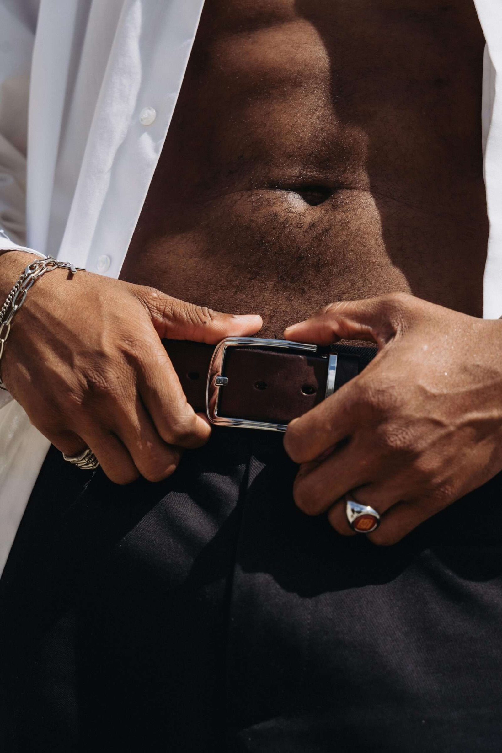 A close-up shot of a man adjusting his leather belt with visible jewelry and an unbuttoned shirt.