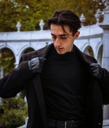 A fashionable man in a black outfit poses confidently outdoors against a classic stone backdrop.