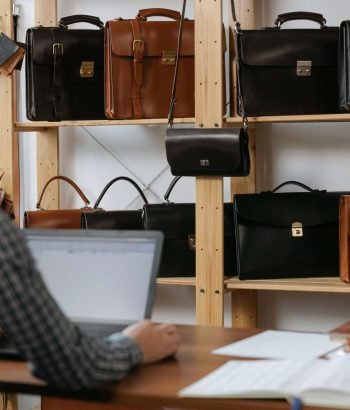 Man at desk using laptop near shelves filled with leather bags.