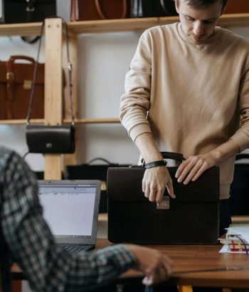 Young artisan selecting leather briefcase in workshop with laptop and numerous bags displayed in the background.